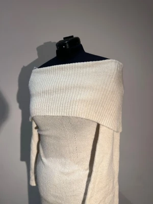 Offshoulder stickad långklänning vit - Superfin vit stickad långklänning med bred offshoulder-krage och långa ärmar. Klänningen har en mjuk och mysig känsla, perfekt för dig som gillar en stilren och trendig look. Passar perfekt till sneakers eller boots.