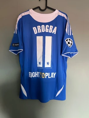 Chelsea Drogba matchtröja blå - Säljer en blå Chelsea matchtröja från Adidas med Drogba och nummer 11 på ryggen. Tröjan har vita detaljer, Champions League-märke på ärmen och texten 'Right To Play' nedtill. Perfekt för dig som älskar fotboll och vill ha en ikonisk supportertröja.