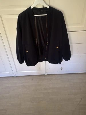 Svart bomberjacka med gulddetaljer - Stilren svart bomberjacka med dragkedja i guld och matchande guldfärgade knappar på fickorna. Jackan har klassisk bombermodell med ribbade muddar och avslappnad passform. Perfekt för dig som vill ha en enkel men snygg jacka till vardags.