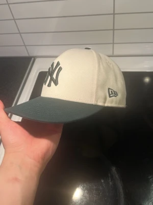 New Era Yankees keps beige/grön - Beige New Era keps med gröna detaljer säljes. Säljer den för har blivit lite för liten, den har ett litet veck på framsidan som ni ser på bild 2 men inget så farligt storlek 7 1/4 57,7 cm