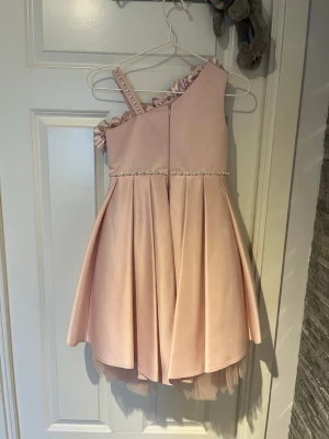 Rosa oneshoulder klänning med volang - Söt ljusrosa kortklänning med oneshoulder och volangdetaljer längs axeln. Klänningen har pärlband i midjan och lager av tyll i kjolen för extra volym. Perfekt för speciella tillfällen och studentfester.
