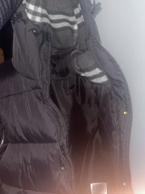 Svart burberry dunväst med huva - Snygg svart dunväst med huva och dragkedja framtill. Västen har quiltad design och är tillverkad i glansigt material. Perfekt för lager-på-lager och passar till många olika outfits.