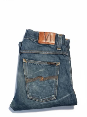 Blå jeans från Nudie Jeans - Snygga blå jeans från Nudie Jeans med klassisk femficksmodell och raka ben. Jeansen har ljusa slitningar och kontrastsömmar samt läderpatch med logga bak i midjan. Tillverkade i bomull för en skön känsla och stilren look.