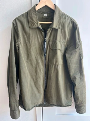 Olivgrön overshirt med dragkedja - Stilren olivgrön overshirt med dragkedja framtill, klassisk krage och bröstficka. Tillverkad i ett mjukt bomullstyg och har en avslappnad passform. Perfekt lager-på-lager-plagg för en cool och avslappnad look.