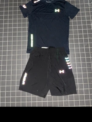 Svart träningsset från Under Armour - Snyggt svart träningsset från Under Armour med t-shirt och shorts. Setet har reflekterande loggor och detaljer i regnbågsfärger. T-shirten har rund hals och korta ärmar, shortsen har resår och dragsko i midjan. Perfekt för gym eller löpning.