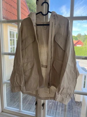Beige jacka från ARKET i linne - Stilren beige jacka från ARKET i italienskt linnetyg från Nova Fides. Jackan har dragkedja, stora fickor framtill och en avslappnad passform. Perfekt för lager-på-lager och snygg till både jeans och kjol.