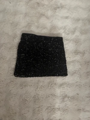 Svart paljettkjol - Snygg svart kortkjol täckt med glittriga paljetter. Perfekt för dig som vill sticka ut och glänsa lite extra. Kjolen har en rak passform och är enkel att matcha med både toppar och skjortor.