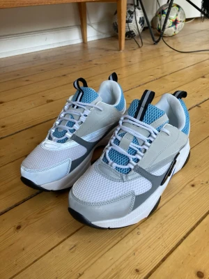 Vita och grå sneakers med blå mesh - Snygga sneakers i vitt och grått med blå meshdetaljer och svarta accenter. Skorna har en chunky sula och vita skosnören. Ovandelen är i mesh och syntetmaterial för en sportig look. Perfekta för dig som gillar streetstyle och bekväma skor.