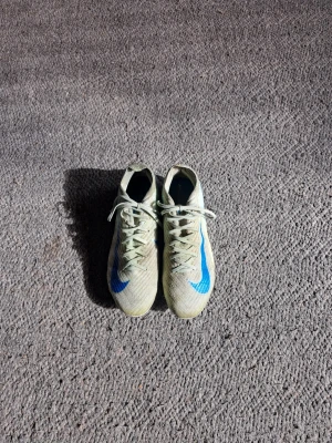 Nike Mercurial fotbollsskor elite - Säljer ett par Nike Mercurial elite! fotbollsskor i beige med blå detaljer och vit sula. Skorna har snörning och är tillverkade i ett lätt syntetmaterial för bra bollkontroll och grepp på planen. Perfekta för dig som vill ha snabbhet och stil på fotbollsplanen. Kostar 3499 ny!