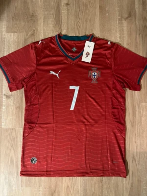 Portugal Ronaldo VM-2026 Fotbollströja - Röd Portugal fotbollströja med nummer 7 och Ronaldo på ryggen. Tröjan har korta ärmar, gröna och röda detaljer vid kragen och är tillverkad i ett lätt, ventilerande material. Perfekt för fotbollsträning eller att bära som supporter. HELT NY