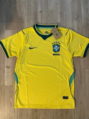 Brasilien VM-2026 Fotbollströja - Gul Brasilien fotbollströja med gröna detaljer och CBF-emblem på bröstet. Tröjan har korta ärmar, V-ringad krage och är tillverkad i ett lätt och ventilerande material. Perfekt för fotbollsträning eller att bära som supporter. HELT NY