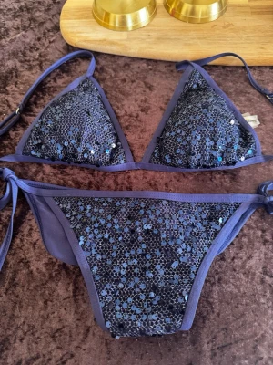 Blå glittrig bikini med paljetter - Snygg blå bikini med glittriga paljetter på både överdel och underdel. Klassisk triangelmodell med knyt i sidorna och runt nacken men även flexibel bikini som kan varieras och ändras till att ha knuten i nacken eller längs axlarna. Perfekt för dig som vill sticka ut på stranden eller vid poolen.