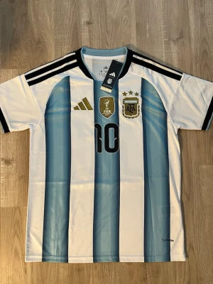 Argentina Messi VM-2026 Fotbollströja - Snygg Argentina matchtröja med Messis namn och nummer 10 på ryggen. Tröjan har klassiska blå och vita ränder, svarta detaljer och guldiga FIFA- och AFA-emblem på bröstet. Perfekt för fotbollsfans som vill visa sitt stöd för landslaget. HELT NY