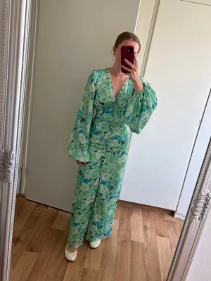 Blommig långklänning från Adoore - Grönblå långklänning från Adoore med blommigt mönster, v-ringning och långa puffärmar. Klänningen har knappar framtill och ett mjukt, flytande tyg som ger en romantisk känsla. Perfekt för dig som gillar färg och mönster. Använt en gång så i ny skick. Nypris 1695kr💞