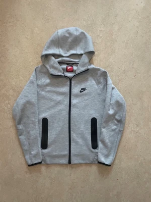 Nike Tech Fleece  - Säljer en klassisk Nike Tech Fleece zip-hoodie i grå med svarta detaljer. Ett av Nikes mest populära och eftertraktade plagg. Har heldragkedja, luva och de karaktäristiska svarta ficköppningarna på framsidan. Nike-loggan broderad på bröstet. Tröjan är 66cm i längden från axlarna ner och 52cm i bredden.