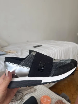 Svarta och silver sneakers från Michael Kors - Snygga sneakers från Michael Kors i svart och silver med elastiskt band över foten och MK-logga. Ovandelen är i textil och syntet med detaljer i silver och svart, samt vit sula. Perfekta för en trendig och sportig look.