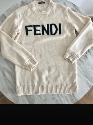 Beige stickad tröja från Fendi - Snygg beige stickad tröja från Fendi med svart FENDI-logga framtill. Tröjan har rund halsringning, långa ärmar och ribbstickade muddar vid ärmslut och nederkant. Perfekt för dig som gillar exklusiva och trendiga plagg.