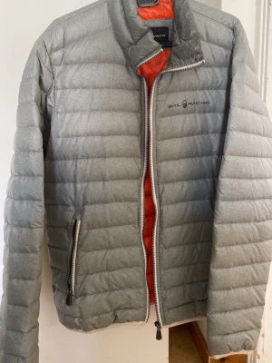Grå dunjacka från Sail Racing - Snygg grå dunjacka från Sail Racing med orange foder och quiltad design. Jackan har två dragkedjeförsedda fickor och hög krage. Perfekt för kalla vinterdagar och har en stilren look med diskret logga på bröstet.