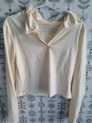 Ljusbeige långärmad topp med krage - En stilren ljusbeige långärmad topp med krage och v-ringning. Toppen har en mjuk och stretchig känsla, perfekt för en trendig och avslappnad look. Passformen är normal och materialet känns som bomull eller syntet.