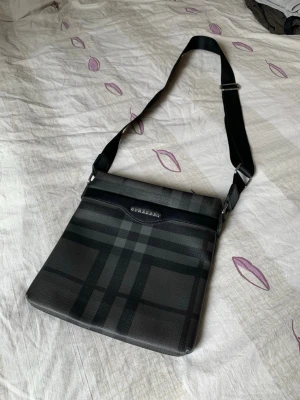Burberry rutig axelväska i svart - Snygg axelväska från Burberry i svart och mörkgrått med klassiskt rutmönster. Väskan har justerbar axelrem i textil, silverfärgade detaljer och en dragkedjeficka på baksidan. Perfekt för dig som vill ha en stilren och praktisk väska med ikonisk design.