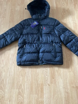  Polo Ralph Lauren jacka - Snygg svart pufferjacka från Polo Ralph Lauren med huva och dragkedja framtill. Jackan har två sidofickor med dragkedja och en bröstficka. Klassisk röd logga broderad på bröstet. Perfekt för kalla vinterdagar.