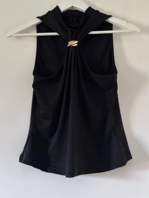 Svart ärmlös topp med gulddetalj - Stilren svart ärmlös topp med snygg drapering framtill och en guldfärgad ringdetalj vid halsen. Toppen har en elegant halterneck-design och är tillverkad i ett mjukt, stretchigt material som ger en smickrande passform.