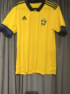 Svenska landslagströja Adidas gul - Gul fotbollströja från Adidas med Sveriges landslagsemblem på bröstet och svarta detaljer. Tröjan har svarta och blå ränder på ärmarna samt Adidas-logga på framsidan. Perfekt för dig som vill stötta Sverige på matchen!