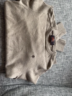 Beige stickad tröja Morris - Beige stickad tröja från Morris med rund halsringning och diskret broderad logga på bröstet. Tröjan har långa ärmar och ribbade muddar vid ärmslut och nederkant. Det står M i den men den är mycket mindre skulle säga en xs då den är för liten för mig
