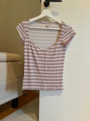 Randig topp med v-ringning - Säljer en ljusrosa topp med svarta ränder och v-ringad hals. Toppen har korta ärmar och är figurnära i modellen. Perfekt att matcha med jeans eller kjol för en avslappnad look.