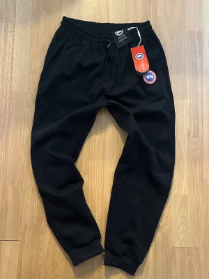Svarta mjukisbyxor från Canada Goose - Svarta mjukisbyxor från Canada Goose med elastisk midja och dragsko. Byxorna har ribbade muddar vid bensluten och en broderad logotyp på ena benet. Perfekta för en chill och avslappnad stil. Materialet är mjukt och bekvämt.