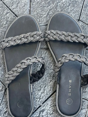 Svarta sandaler med glittriga flätade band - Snygga svarta sandaler från ENVIE med två breda flätade band täckta av små glittrande stenar. Sandalerna har en platt sula och dekorativa stenar längs kanten för extra bling. Perfekta för att lyfta vilken outfit som helst.