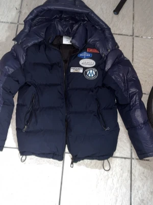 Mörkblå dunjacka från Mercier - Snygg mörkblå dunjacka från Mercier med flera coola patches på bröstet. Jackan har dragkedja framtill, två sidofickor med dragkedja och en hög krage med tryckknappar. Perfekt för kalla vinterdagar och riktigt trendig look. På bild 3 ser man att lite av dragkedjan gått av, men att det däremot går att knäppa jackan 