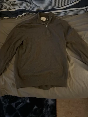 Grå långärmad tröja med half zip - En stilren grå långärmad tröja med half zip och hög krage. Perfekt för lager-på-lager och enkel att matcha med jeans eller chinos. Tröjan har ribbade muddar och är tillverkad i ett mjukt material som känns skönt mot huden. Ifrån bläck.