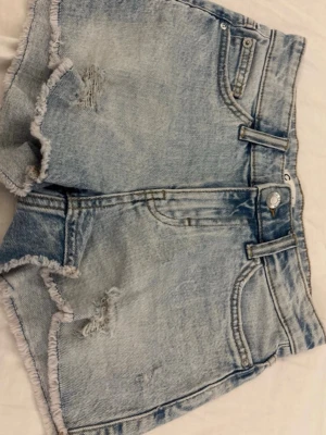 Ljusblå jeansshorts  - Snygga ljusblå jeansshorts med fransiga kanter och slitna detaljer framtill och baktill. Klassisk femficksmodell med bälteshällor och knappstängning. Perfekta för varma dagar och en avslappnad stil.