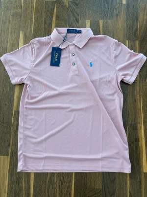 Ljusrosa pikétröja Ralph Lauren - Snygg ljusrosa pikétröja från Polo Ralph Lauren med klassisk krage, korta ärmar och två knappar framtill. Tröjan har den ikoniska blå logotypen broderad på bröstet och är tillverkad i ett mjukt bomullsmaterial. Perfekt för en clean och stilren look.