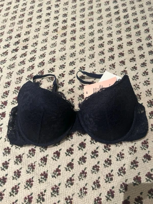Mörkblå push up-bh från H&M - Snygg mörkblå push up-bh från H&M med spetsdetaljer och vadderade kupor. Justerbara axelband och klassisk knäppning bak. Perfekt för dig som vill ha extra lyft och en feminin look.