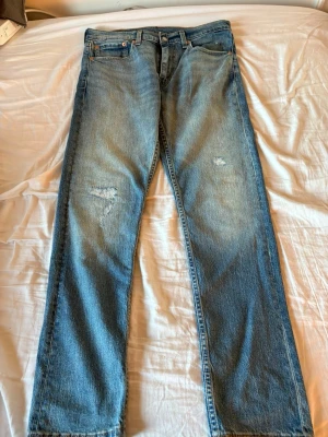Blå raka Levis jeans W33 L32 - Säljer ett par klassiska blå Levis jeans  med raka ben och lätt slitna detaljer på låret. Jeansen har fem fickor och normal midja. Perfekta för en avslappnad och trendig look.