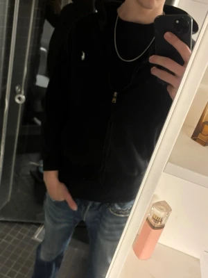Svart hoodie från Polo Ralph Lauren - Klassisk svart hoodie från Polo Ralph Lauren med dragkedja framtill och vit broderad logga på bröstet. Hoodien har huva, känguruficka och ribbade muddar vid ärmslut och nederkant. Tillverkad i mjuk bomullsblandning för skön komfort. Säljer pga för liten storlek