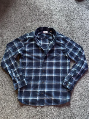 Rutig Skjorta - Snygg blå rutig skjorta med långa ärmar och klassisk krage. Skjortan har knäppning framtill och vid ärmsluten. Perfekt för en avslappnad och stilren look. Storlek L men sitter mer som M!