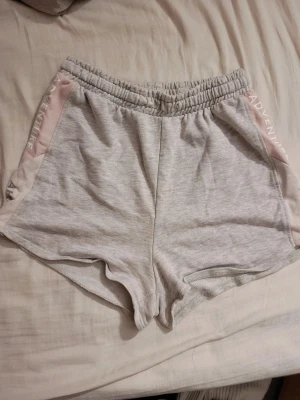 Grå sweatshorts med rosa detaljer - Mysiga grå sweatshorts med elastisk midja och ljusrosa paneler på sidorna där det står 'ADVENTURE' tryckt i vitt. Perfekta för chill dagar eller träning. Mjukt material och avslappnad passform.