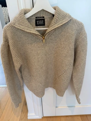 Exklusiv stickad tröja från Scotch & Soda Amsterdam i storlek 164 (13–14 år) säljes i kanonskick. - Elegant melerad beige nyans kombinerad med en sofistikerad half-zip i guldfärg som ger plagget en lyxig och modern känsla. Tröjan har vackra stickade detaljer, avslappnad passform med nedsänkta axlar samt en mjuk och värmande garnblandning med ull och mohair för extra komfort.  • Storlek 164 / 13–14 år • Kanonskick • Djur- och rökfritt hem  Ett stilrent premiumplagg som passar perfekt för både vardag och mer uppklädda tillfällen. 🤍