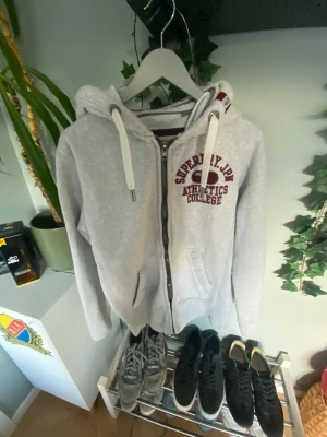 Superdry hoodie - Snygg ljusgrå hoodie från Superdry med dragkedja framtill och tryck i vinrött på bröstet. Utrustad med huva och vita dragsnören. Perfekt för en avslappnad och sportig stil. Mjuk insida och klassisk collegekänsla.