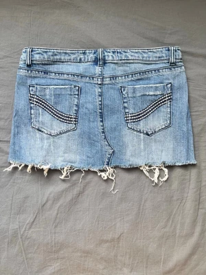 Blå jeanskjol med fransig kant - Säljer en blå jeanskjol med slitna detaljer och fransig kant. Kjolen har fem fickor, bälteshällor och dekorativa sömmar på bakfickorna. Perfekt för dig som gillar en avslappnad och trendig stil. Midjemått 80 cm, längd 25/30 cm