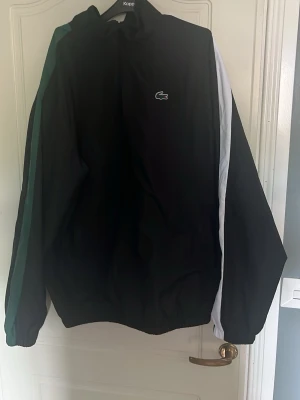 Svart Lacoste tracksuit  - Snygg svart vindjacka från Lacoste med gröna och vita ränder längs ärmarna. Jackan har dragkedja, hög krage och elastiska muddar. Klassisk Lacoste-logga på bröstet och stor logga med krokodil på ryggen. Perfekt för en sportig och trendig stil. Strl Xxl men passar perfekt till dig som är XL i storlek Priset går att diskutera    Koftan eller byxorna kan köpas separat 