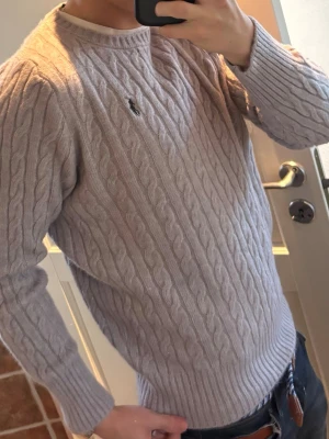 Ljusgrå kabelstickad tröja Polo Ralph Lauren - Nästan helt ny, fick denna i julklapp! Kabelstickad ljusgrå tröja från Polo Ralph Lauren med rund halsringning och broderad logga på bröstet. Tröjan har långa ärmar och ribbade muddar, perfekt för en stilren och klassisk look. 