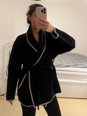 Svart kappa med vita detaljer - Snygg svart omlottkappa med vita sicksack-detaljer längs kanterna. Kappan har ett brett skärp i midjan och avslappnad passform. Materialet är ullblandning. Använd ett fåtal gånger och i nyskick. Nypris 500kr. Lite större i storlek. Passar en M och L. Från Kappahl.