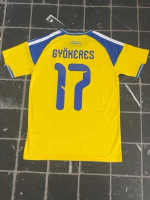 Sverige fotbollströja Gyökeres 17 - Gul och blå fotbollströja från Adidas med Sveriges landslagsemblem på bröstet och Gyökeres samt nummer 17 på ryggen. Tröjan har korta ärmar, blå detaljer på axlar och ärmslut samt klassiska Adidas-ränder. Perfekt för dig som vill stötta Sverige på match!