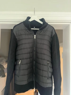 Svart cardigan med dragkedja - Snygg svart cardiganjacka från by Aldén med och stickade ärmar. Säljer den pågrund av att storleken inte passade. Den är helt oanvänd och har förpackning och allt annat kvar. Jackan har två fickor och hög krage. Perfekt för lager-på-lager och ger en sportig men ändå stilren look.