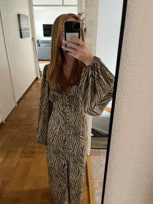 Zebramönstrad långklänning från Adoore - Snygg långklänning från Adoore med zebramönster i beige och brunt. Klänningen har långa, vida ärmar, v-ringning och knappar framtill.  Jättefint skick! Jag är 160 cm och den går precis ner till marken 