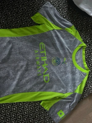 Manchester City tränings t-shirt Puma - Grå och neongul Manchester City tränings t-shirt från Puma med Etihad Airways-logga på bröstet. T-shirten har sportig design, rund halsringning och detaljer i neonfärg längs sidor och ärmar. Perfekt för fotbollsträning eller gym.